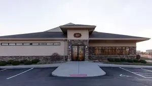 An exterior view of a building with a gray roof, brick walls, and glass windows, situated in an empty parking lot with bushes and a sidewalk