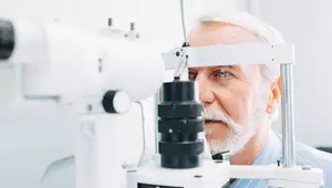 An elderly man with white hair and a beard is having his eyes examined by an optometrist using an ophthalmoscope.