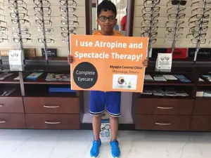 A young boy is holding a sign that reads 'I use Atropine and Spectacle Therapy!' and is standing in front of an optometrist's office.