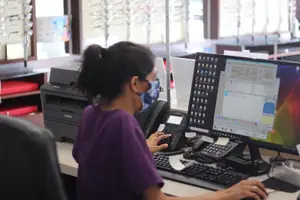 A woman wearing a mask is sitting in front of a desk in an office with a computer monitor, keyboard, and telephone