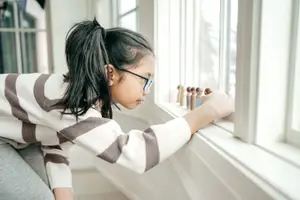A girl with glasses looking outside through a window with wooden figurines on the sill.