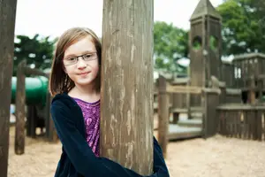 A girl in glasses stands in a playground, holding a wooden pole, with a slide and tower in the background.