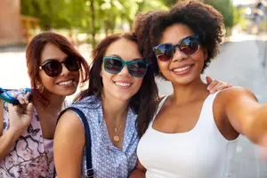 Three women in sunglasses smiling and posing for a selfie on a sidewalk