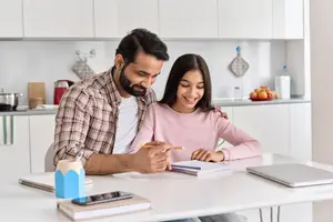 A man and a girl sitting at a table in a kitchen, looking at a notebook, smiling, and having a conversation