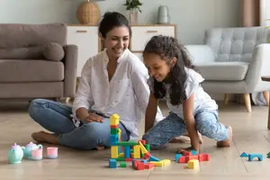 A woman and a girl are playing with colorful building blocks on the floor, with a couch and a lamp in the background.