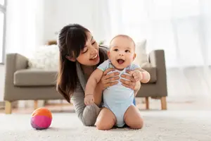 A woman smiling and holding a baby on the floor with a colorful ball nearby.