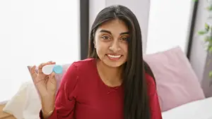 A smiling woman in a red shirt is sitting on a bed and holding a blue and white contact lens.