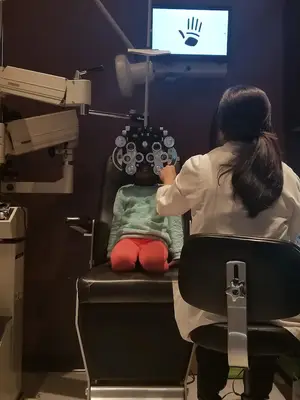 A girl in a green sweater is sitting on a chair while a woman in a white lab coat is doing an eye exam on her in a clinic