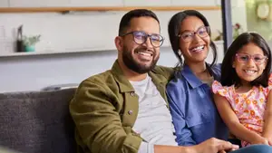 A family of three sitting on a couch, smiling, with a girl in a pink floral dress and glasses, a man in a green jacket, and a woman in a blue shirt, and a cabinet with a potted plant behind them.