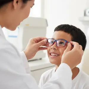 A young boy with blue frames is smiling as an adult female optometrist adjusts his glasses.