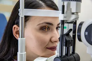 A woman with brown hair is looking at something through a medical eye examination machine, her right eye is slightly open, and her left eye is closed, with a bright light illuminating her eye.