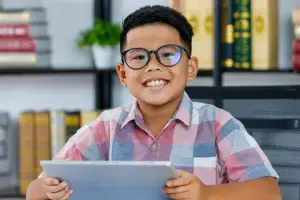 A smiling boy with glasses is holding a tablet in a room with bookshelves and a plant in the background.