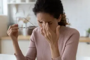 A woman in a pink sweater is sitting in a kitchen and holding her glasses while covering her face with her hands.
