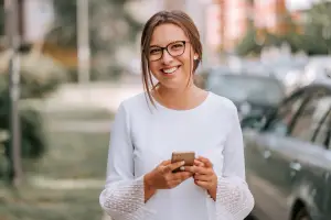 A woman wearing eyeglasses is standing and smiling on the side of the road.