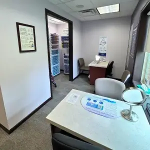 A picture of a room that has a desk with a mirror and a sign on it, a chair, a table, and a cabinet with several drawers, and a window with a frame and a blind.