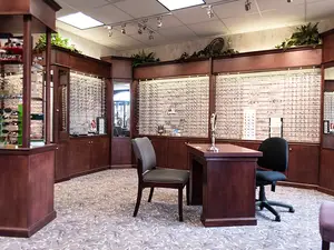 An office with a desk, chairs, and a glass cabinet filled with glasses, set against a backdrop of a white wall adorned with mounted frames, illuminated by ceiling lights, and featuring a mirror on the right side, all contributing to a professional and organized atmosphere.