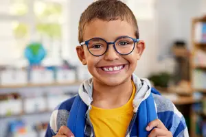 A smiling boy with glasses and a backpack is in a classroom with shelves and a globe in the background.