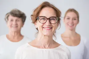 Three women standing together and smiling at the camera, one of them wearing glasses