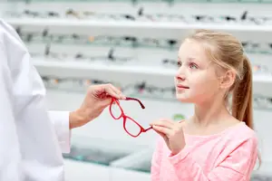 A young girl is holding a pair of red glasses in her hands while looking at them in a store