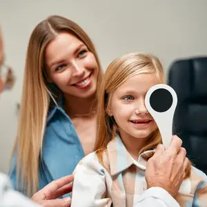 A young girl is having her eyes examined by a doctor in a clinic, with her mother standing beside her, all smiling.