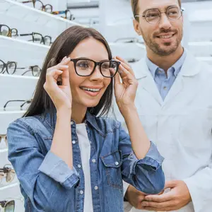 A smiling woman trying on glasses in a store with a smiling man in a white coat behind her