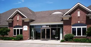 A modern brick building with a gray roof, large windows, and a glass entrance displaying the name 'Pro Care', surrounded by greenery and trees.