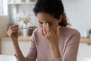 A woman sits at a table holding a pair of glasses, touching her forehead, and looking tired