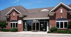 A brick building with a gray roof, glass doors, and windows, and plants in front, with a clear blue sky above.