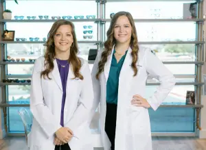 Two women in white lab coats standing in front of a shelf with glasses