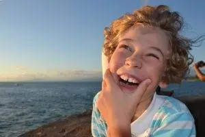 A young boy is smiling and seems to be posing for a photo with a hand on his cheek while standing on a rocky shore near the sea during sunset