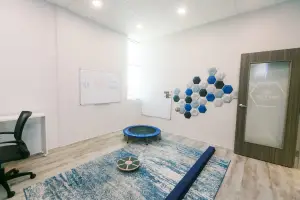 A well-lit indoor playroom with a blue carpet, a desk, a chair, a trampoline, a whiteboard, and a glass door.