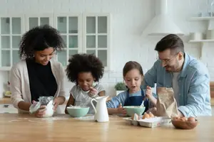 Four people are in the kitchen, a woman is pouring flour into a bowl, a girl is looking at something, a boy is holding a spoon, and a man is standing beside the boy.