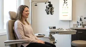 A woman is sitting in an eye exam chair while smiling at the camera
