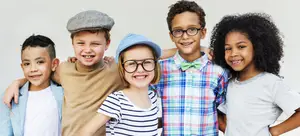 Five children smiling and posing for a photo with two wearing glasses and one with a hat