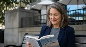 An older woman in a gray shirt and blue blazer sits on a bench outdoors and reads the book Clear Horizons