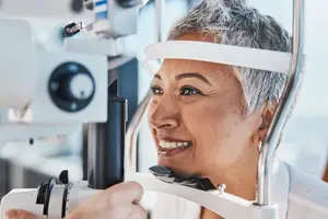 A woman with gray hair and a white headband smiles while being examined by an eye doctor using a device.