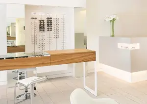 A modern, white-walled optical shop with a wooden counter, a white chair, and a glass display of sunglasses, accompanied by a white flower in a green vase on the wall, creating a clean and inviting atmosphere.