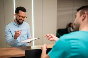 A man wearing glasses is smiling and holding a pen and clipboard in his hand while talking to another man in a blue shirt in a room