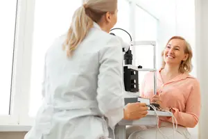 Woman with pink top smiling with doctor holding eye exam equipment
