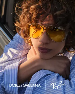 A woman with curly hair wearing blue and white striped shirt and yellow Ray-Ban sunglasses sits in a car.