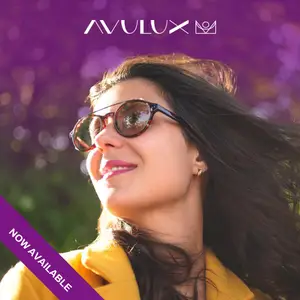 A smiling woman with dark hair wearing sunglasses and a yellow jacket poses for a photo against a purple background.