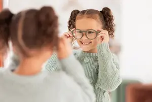 A girl wearing glasses is looking at herself in a mirror while a woman holds the glasses for her.