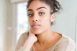 Close-up of a woman with curly hair, light brown skin, and blue eyes, looking thoughtful and slightly touching her chin with her right hand, wearing a beige sweater in a room with white walls and a window in the background.