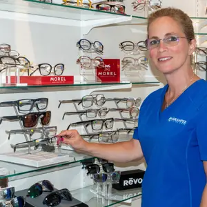 A woman with glasses is standing in front of a shelf full of glasses and sunglasses. She is wearing a blue shirt with the word Mashpee on it. She is holding a pair of glasses and smiling.