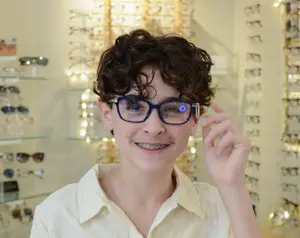 A young person with braces wearing glasses and smiling in front of a display of glasses