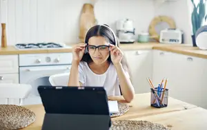 A woman wearing glasses and headphones is sitting at a table with a laptop in a kitchen.