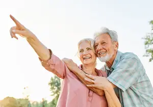 A smiling elderly couple standing together in a park with trees in the background and a clear sky