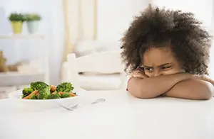 A young girl sitting at a table with a plate of broccoli and carrots, looking upset.