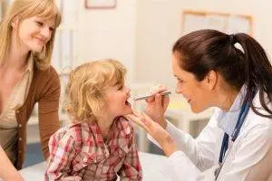 A doctor uses a stethoscope to examine a child who is blowing into a spirometer while a woman watches. Behind them, there is a wall with pictures hanging on it.
