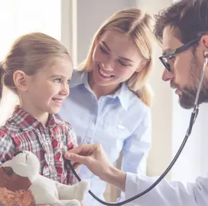 A doctor is examining a child with a stethoscope while the child's mother looks on, all three smiling at the camera.